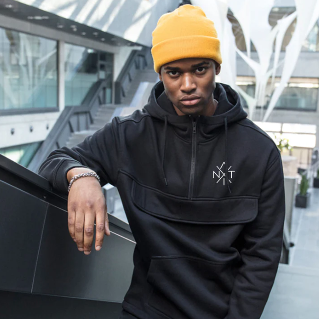 Person wearing a black hoodie with a logo, yellow beanie, and dark pants in an urban setting.
