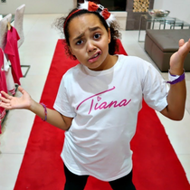 Child wearing a 'Tiana' shirt in a room with red and pink shirts on a rack.